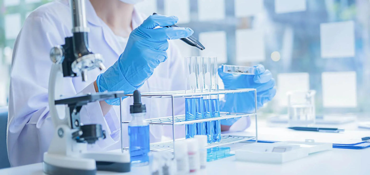 Scientist wearing a lab coat and blue gloves using a dropper to fill test tubes with IGF-1 liquid in a laboratory, with a microscope and various lab equipment in the foreground.