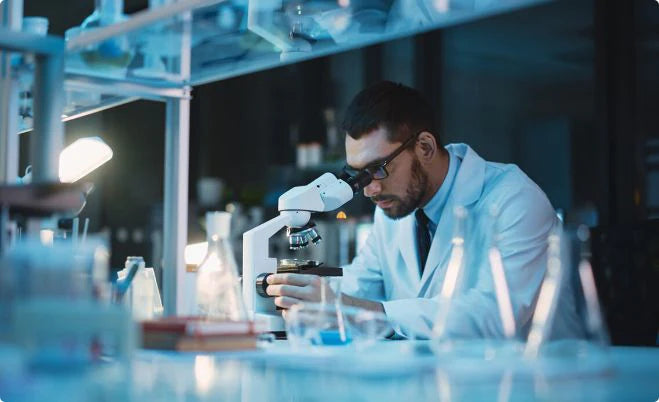 Scientist in a lab coat using a microscope to study deer antler velvet, surrounded by lab equipment and beakers under dim, focused lighting.