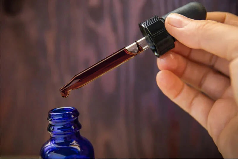 Hand holding a dropper filled with IGF-1 Plus liquid above a blue glass bottle, set against a blurred wooden background.