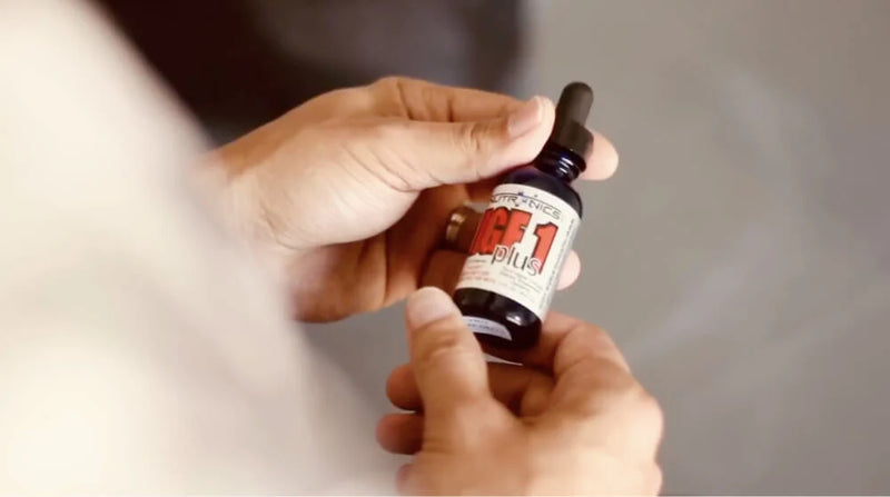 Close-up of hands holding a bottle of Nutronics Labs IGF-1 Plus supplement, focusing on the product label and dropper cap.