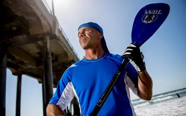 A middle-aged man stands confidently near a beach, holding a blue paddle in one hand. He is wearing a bright blue athletic shirt with white accents and a blue cap. The background features a pier extending over the water, with clear skies and gentle waves visible. The man has a serious expression, looking slightly off to the side.